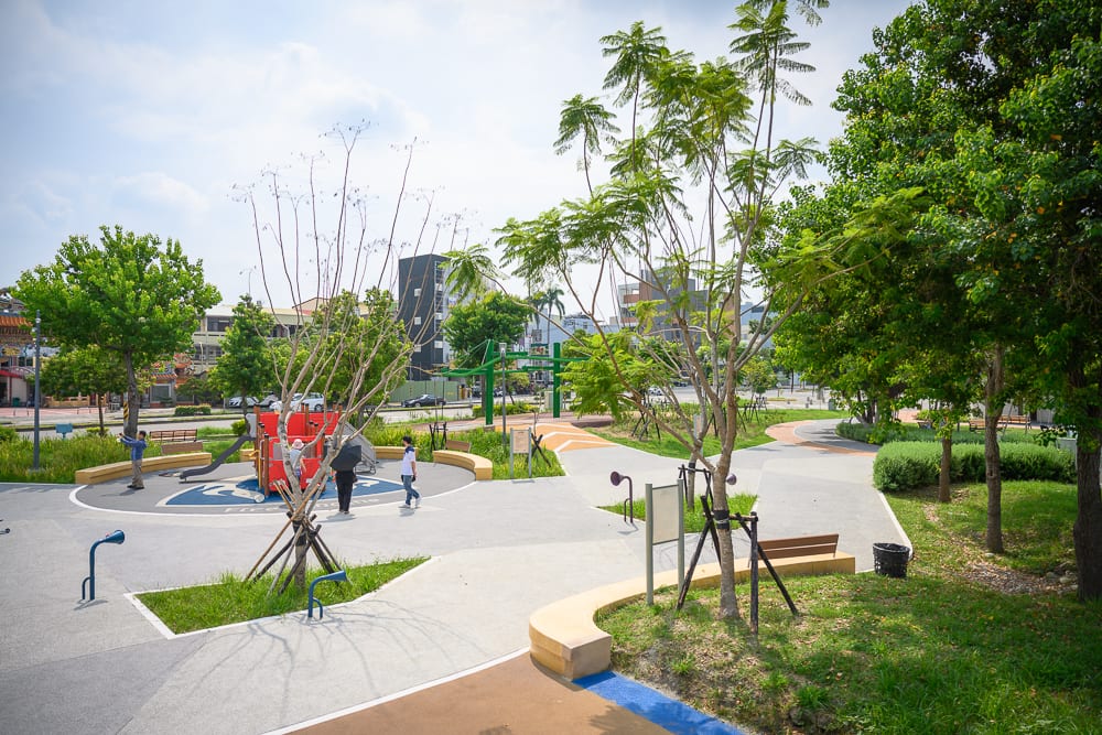 彰化員林兔馬鹿公園完整指南|親子遊戲場玩法解析 5 彰化兔馬鹿公園|綠樹、公園、遊樂設施、人行步道、座椅,日間戶外開放空間鋪設灰色地磚與草皮,遠處有低矮建築與道路車輛。