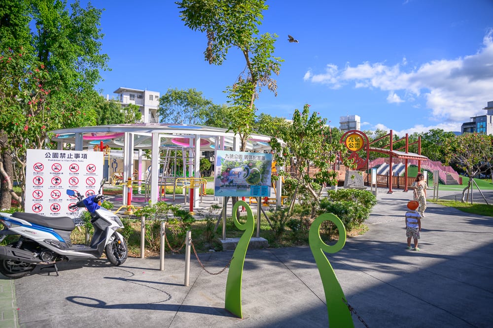 宜蘭九號公園｜機車、公園、涼亭、告示牌、綠樹、藍天白雲、行人。白色機車停在公告牌旁，露天廣場設有白色圓柱涼亭與遊樂