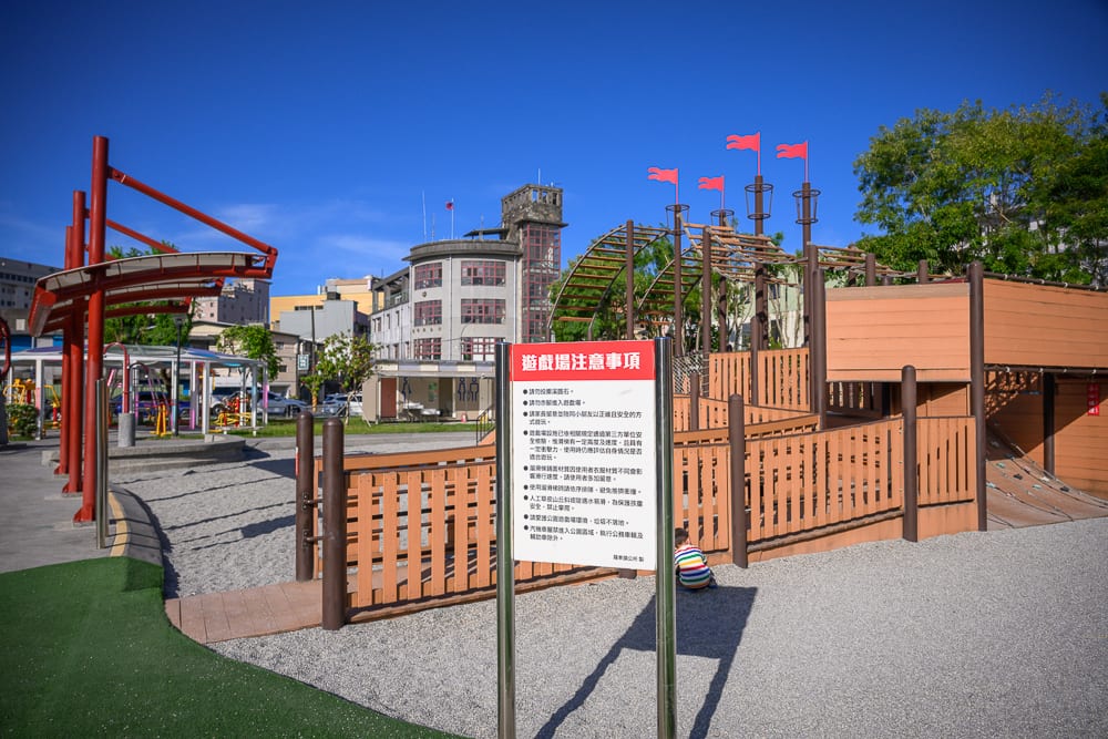 宜蘭九號公園｜藍天下的戶外公園，遊樂場，木造船型遊具，告示牌，紅色旗幟，沙地，孩童，灰色建築物，鐵架。