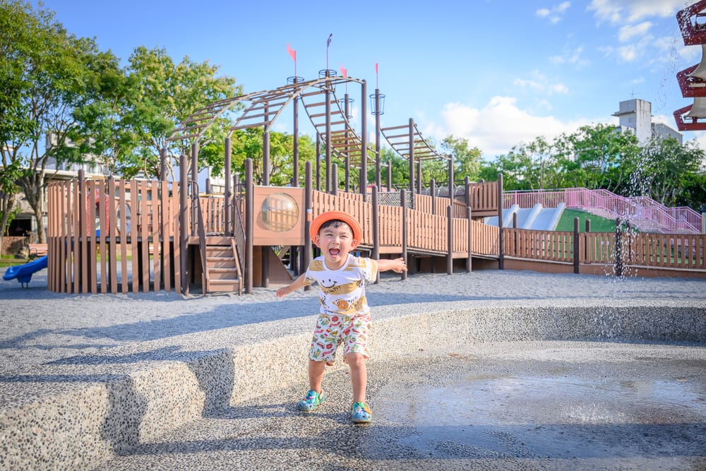 宜蘭九號公園｜兒童遊樂場、男孩、水池、木製遊具、沙地、藍天，男孩戴著橘色帽子張嘴大笑，站在濕漉漉的水泥地噴水設施旁