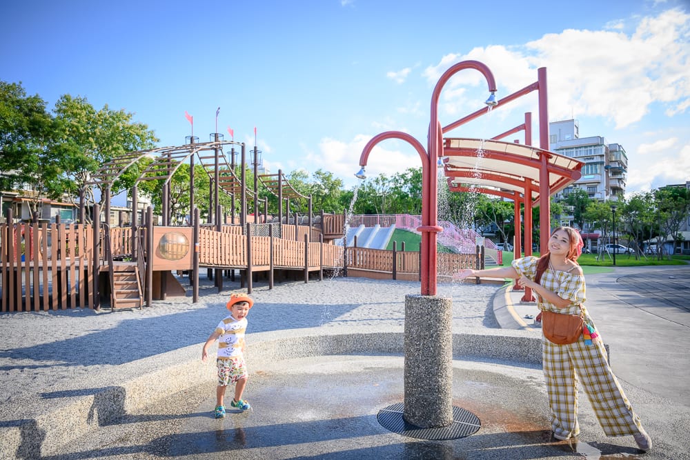 宜蘭九號公園｜戶外遊樂場、噴水柱、兒童、女性、沙坑、木造遊具、藍天、白雲。穿格紋衣的女性站在紅色噴水柱旁，幼童在水