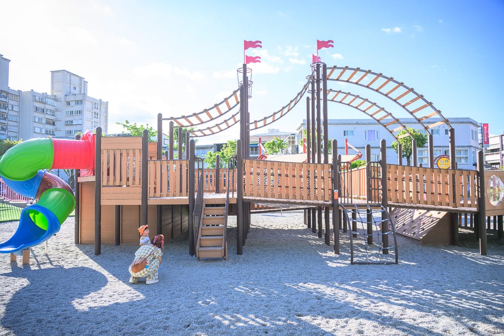 宜蘭九號公園｜戶外公園、遊樂場、大型木製遊具、攀爬架、紅綠藍三色螺旋滑梯、沙地、大人與小孩、背景大樓與藍天白雲。
