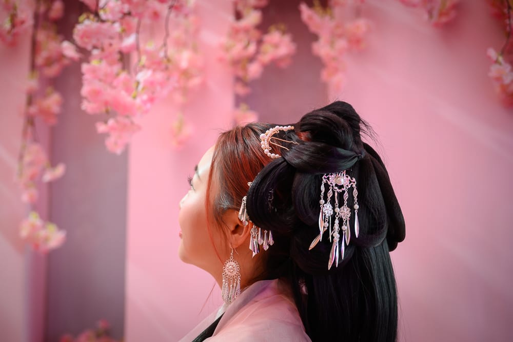 台中RJ55漢服完整指南|漢服服飾與古風配件 12 台中RJ55漢服|女性側臉,古典盤髮,金屬髮簪,耳環,粉紅牆面,人工櫻花枝,室內,粉紅光線。