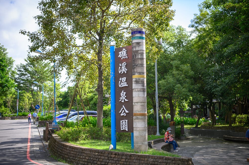 宜蘭礁溪溫泉公園完整指南|兔子裝置藝術、涼亭與步道 9 宜蘭礁溪溫泉公園|公園,招牌,樹木,路燈,行人。寫著「礁溪溫泉公園」直立招牌立於綠地中央,旁側有圓柱造景。兩側為綠樹與石磚步道,遠處可見車輛停放,行人散步。