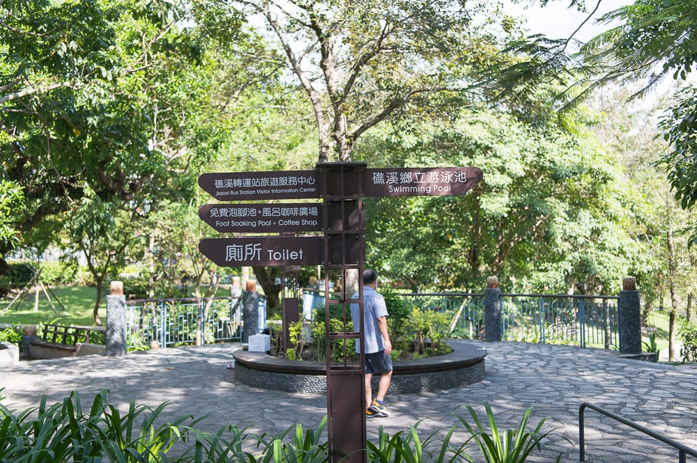 宜蘭礁溪溫泉公園完整指南|兔子裝置藝術、涼亭與步道 5 宜蘭礁溪溫泉公園|指標、廣場、樹木、欄杆、行人,樹林間立著棕色雙向指標牌,指引方向涵蓋游泳池與洗手間。鋪面石板廣場旁有圓形花圃與護欄,穿短袖短褲的男子背對鏡頭站立。