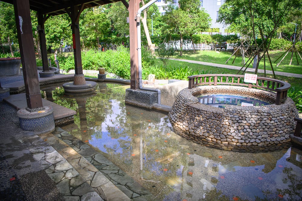 宜蘭礁溪溫泉公園完整指南|兔子裝置藝術、涼亭與步道 4 宜蘭礁溪溫泉公園|木造涼亭、石砌水池、矮圍欄、水面反射樹影、戶外公園、綠地草皮、排水管、石板步道。涼亭木柱支撐屋頂,池水清澈映出天空與綠樹,右側有圓形石砌造景帶有木製圍欄。