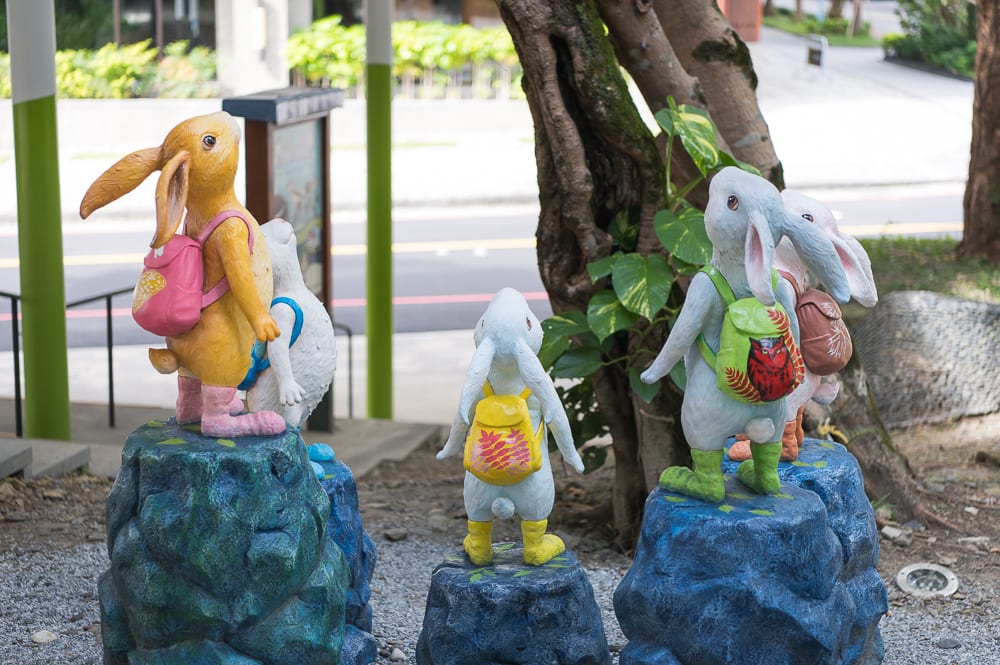 宜蘭礁溪溫泉公園完整指南|兔子裝置藝術、涼亭與步道 3 宜蘭礁溪溫泉公園|兔子雕像、背包、藍色石塊、樹木、街道,戶外公共藝術裝置,數隻背著彩色背包的兔子雕像站立在藍色石座上,背對觀者面向道路。