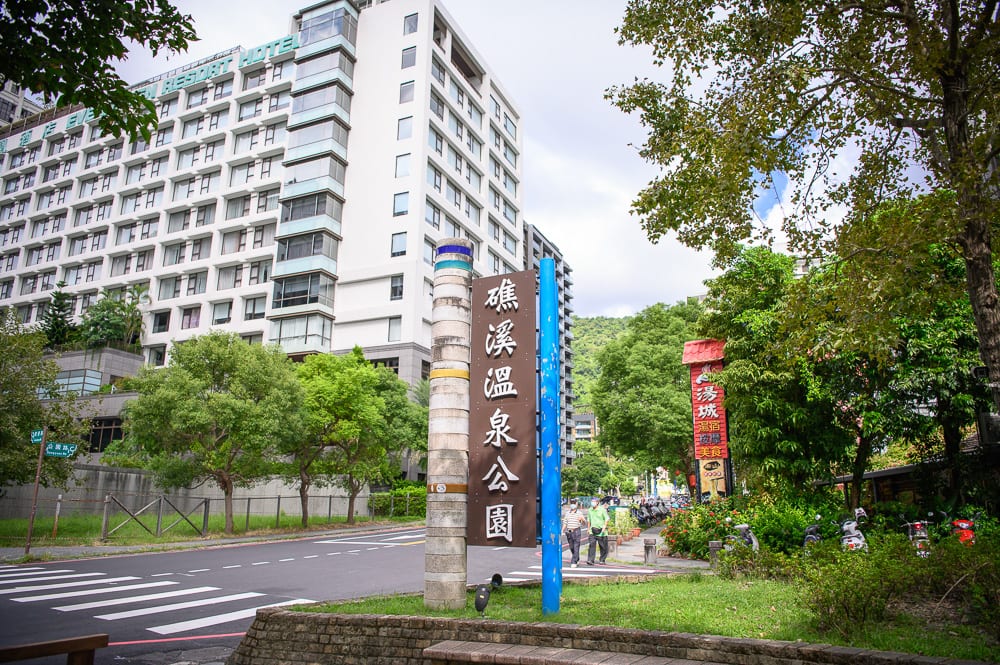 宜蘭礁溪溫泉公園完整指南|兔子裝置藝術、涼亭與步道 6 宜蘭礁溪溫泉公園|街道、飯店、公園標示牌、綠樹,白色高聳大樓與灰色柏油路面並置,公園立牌寫著礁溪溫泉公園,路旁停放數輛機車。