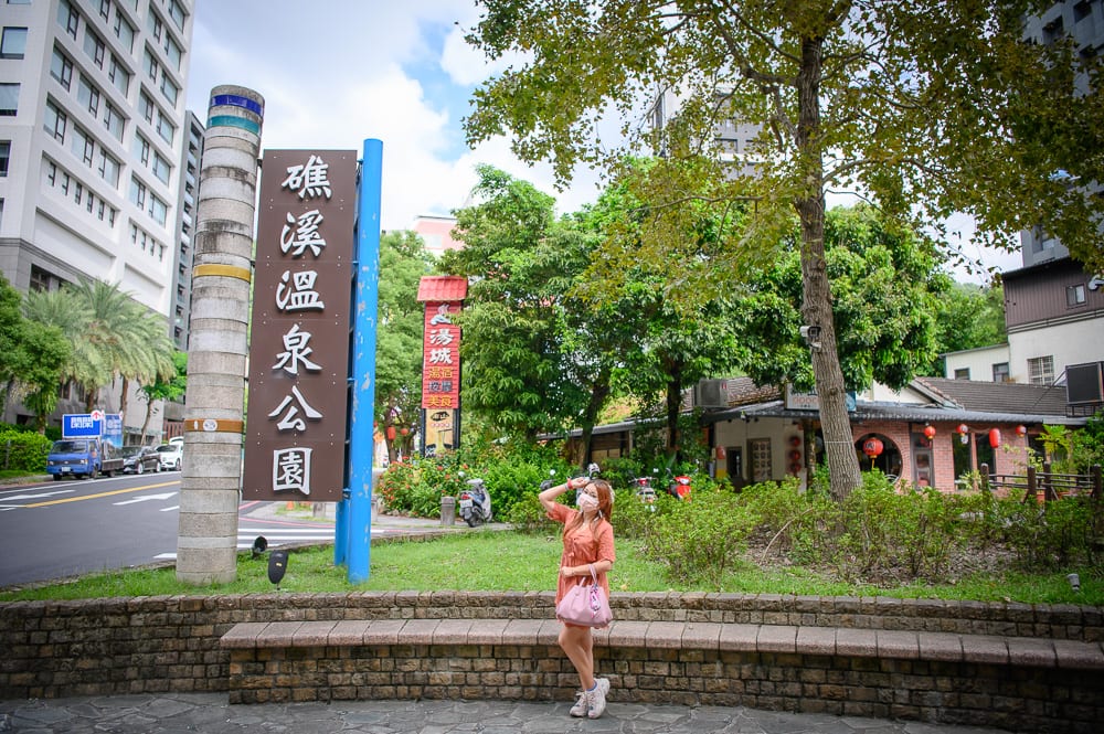 宜蘭礁溪溫泉公園完整指南|兔子裝置藝術、涼亭與步道 8 宜蘭礁溪溫泉公園|街道旁,女子,招牌,樹木,建築,石牆,公園。一名戴口罩的女子站在「礁溪溫泉公園」大型直立木製招牌旁,前方為馬路與高樓建築,後方有綠樹與低矮平房,地面為石磚鋪面與矮牆。