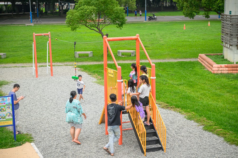 彰化景觀公園完整指南|兒童遊樂設施與高架橋下綠地玩法 12 彰化景觀公園|公園、草地、溜滑梯、遊具、兒童、成人,大人與小孩聚集在橘色金屬遊具周圍,地面鋪滿碎石,背景有大片綠地
