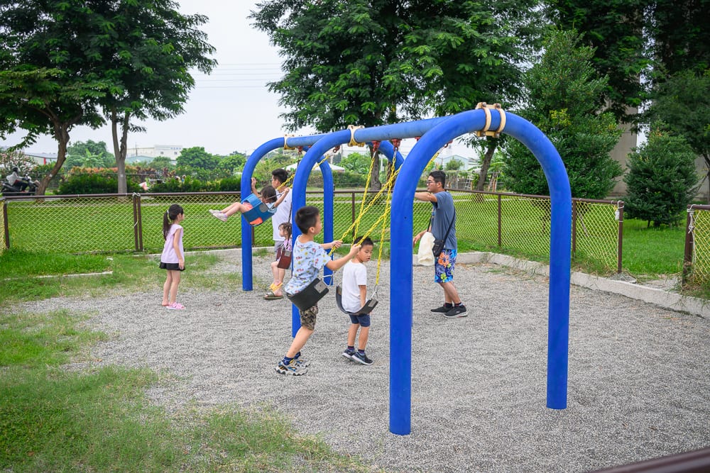 彰化景觀公園完整指南|兒童遊樂設施與高架橋下綠地玩法 11 彰化景觀公園|公園、藍色鞦韆、兒童、成年人、碎石地、草地、樹木,大人與小孩在戶外遊樂場互動,金屬管架支撐著吊繩與座