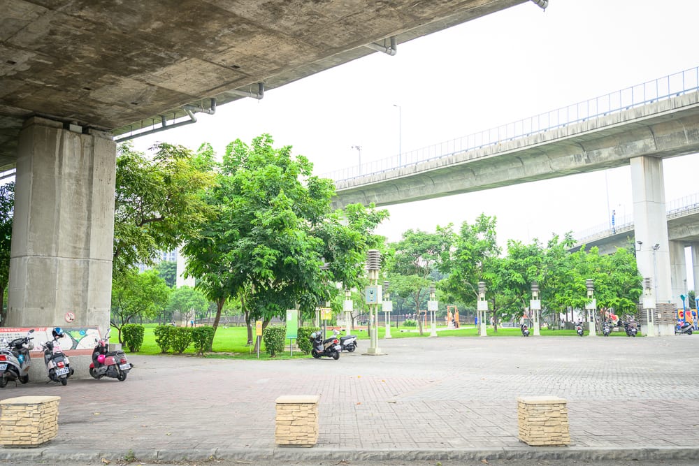 彰化景觀公園完整指南|兒童遊樂設施與高架橋下綠地玩法 8 彰化景觀公園|高架橋墩、水泥橋樑、綠樹、草皮、廣場、機車、石椅,灰白天空與陰影覆蓋的高架橋下空間,地面鋪設磁磚。