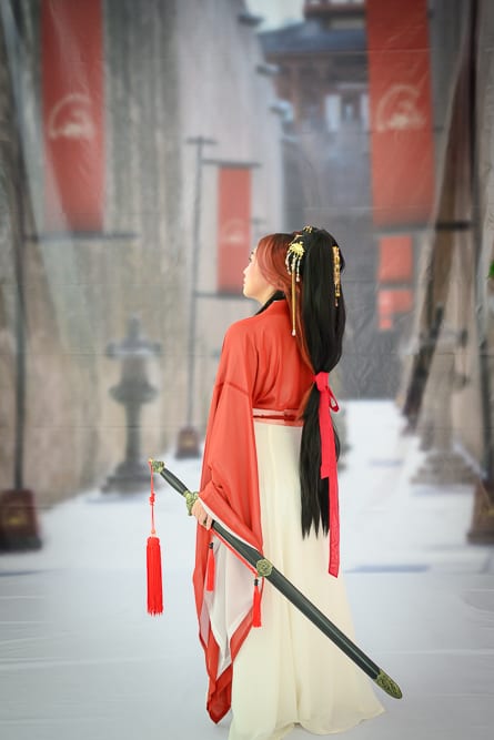 台中RJ55漢服完整指南|漢服服飾與古風配件 2 台中RJ55漢服|女性、古裝、長劍、紅色布旗、石造建築、長廊。女性背對鏡頭站立,身穿紅白色古裝長袍,梳著高馬尾髮髻並插有金色髮簪,右手持一把帶有紅色流蘇的長劍。背景為延伸的石造走廊,兩側懸掛紅色直式布旗。