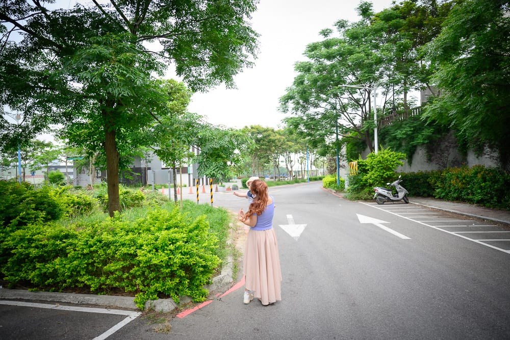 新竹芎林水霧森林遊戲場完整指南|幼兒遊戲設施 6 新竹芎林水霧森林遊戲場|街道、綠樹、灌木叢、背影、女子、機車、白色箭頭標線。長髮女子手抱幼童站在柏油路旁,兩側茂密枝葉與翠綠草皮延伸至遠方,地面繪有指引箭頭與停車格線。