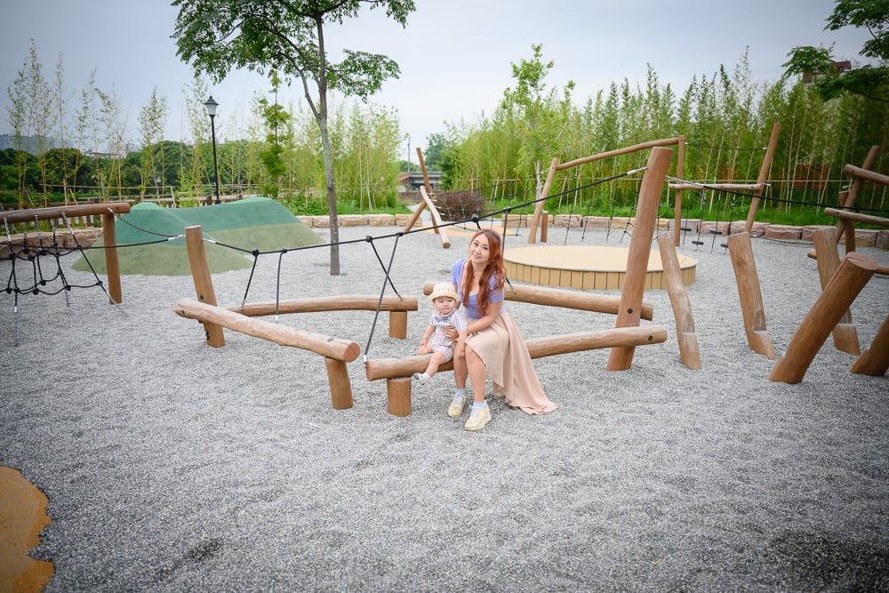 新竹芎林水霧森林遊戲場完整指南|幼兒遊戲設施 12 新竹芎林水霧森林遊戲場|女性與幼兒、戶外遊樂場、圓木攀爬架、灰色碎石地、竹林背景。女性坐在木樁上抱著戴帽子的幼兒,周圍有木頭欄杆、繩網設施與綠色草皮山丘。