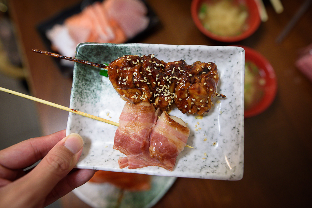 台北三味食堂｜手持餐盤，上方擺放日式烤雞肉串與培根捲，背景為餐廳桌面。