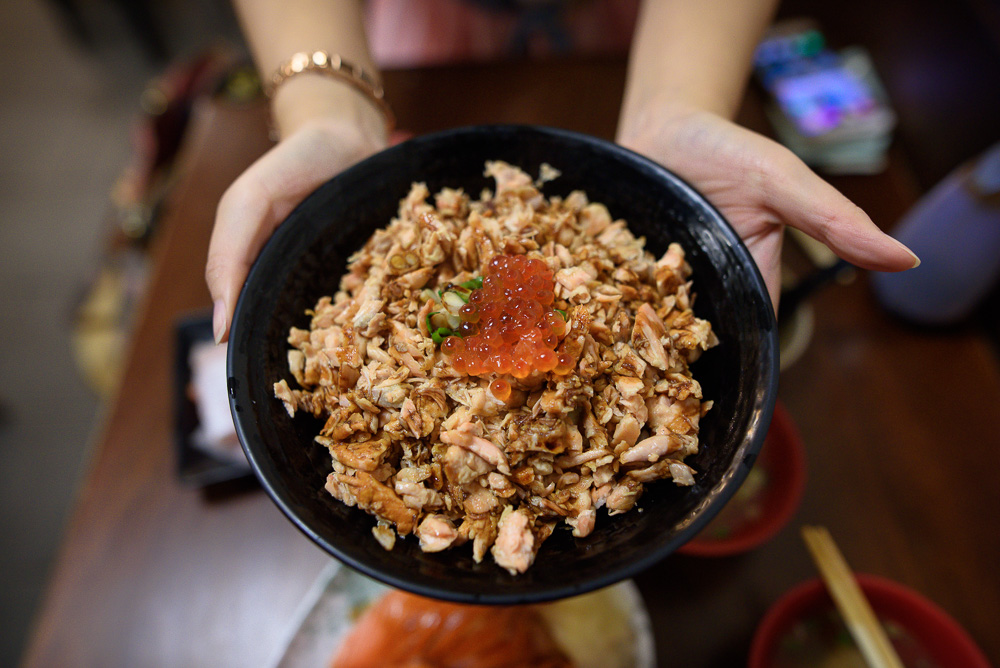 台北三味食堂｜雙手捧著盛滿碎鮭魚與鮭魚卵的黑色碗，室內餐廳桌面。