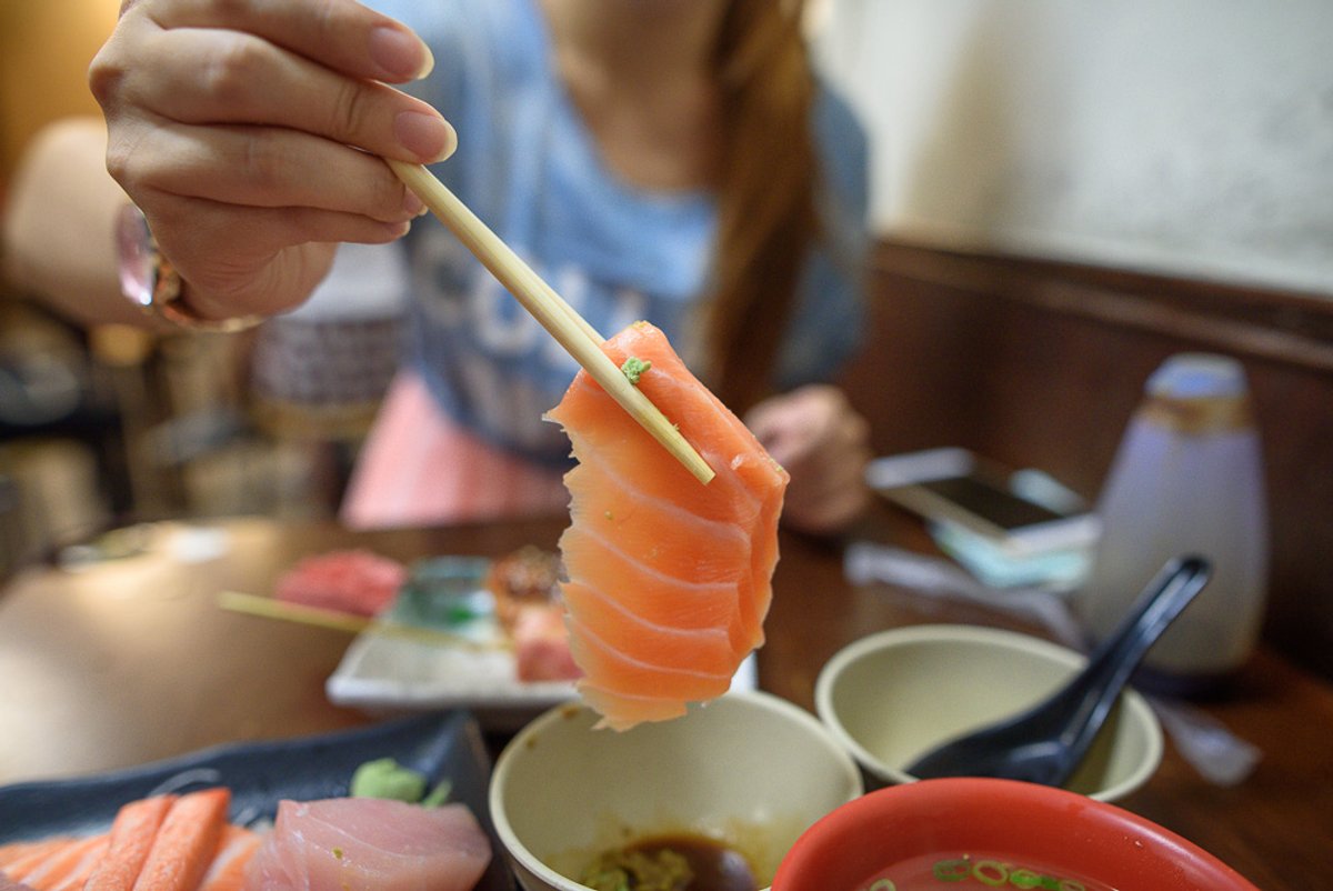 台北三味食堂｜餐廳內，手持筷子夾起一片鮭魚生魚片，桌上有醬料碗、湯碗與其他餐點。