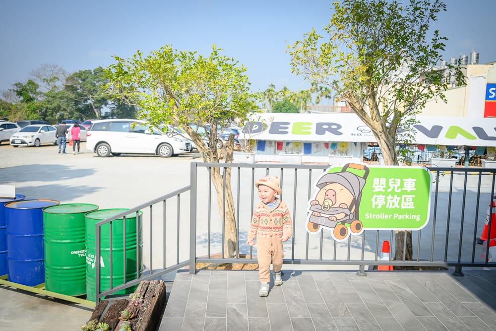 南投鹿芝谷生態景觀園區完整指南|餵羊、水豚與親子玩法 7 南投鹿芝谷|戶外廣場、幼童、鐵欄杆、嬰兒車停放區告示牌、油漆桶、樹木、白色汽車、遠方建築物,晴朗天空下地面平坦,彩色圓桶排列於樓梯旁。