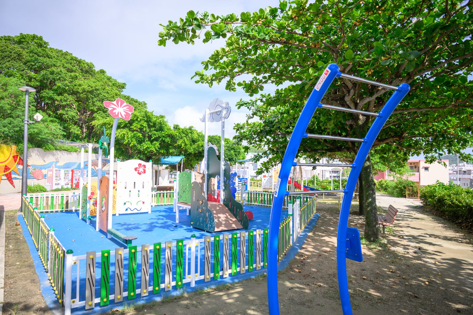 日本沖繩海軍壕公園完整指南|地下壕博物館與兒童遊樂場 10 日本沖繩海軍壕公園|戶外公園,藍色防墜地墊,溜滑梯,彩色欄杆,藍色攀爬架,綠樹,步道,長椅,遠方建築。