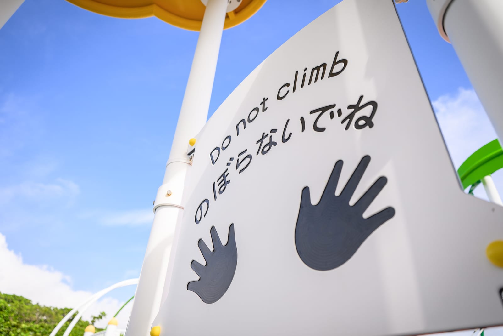 日本沖繩海軍壕公園完整指南|地下壕博物館與兒童遊樂場 7 日本沖繩海軍壕公園|標示牌、遊具、天空、英文、日文字幕,白色面板印有雙手圖案與禁止攀爬字樣,搭配藍天背景。