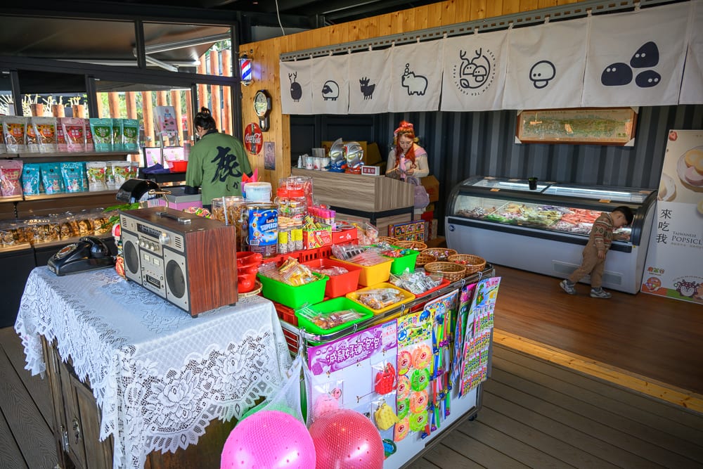南投鹿芝谷生態景觀園區完整指南|餵羊、水豚與親子玩法 5 南投鹿芝谷|室內,復古雜貨店。櫃檯陳列錄音機、零食罐、彩色塑膠籃,前方鋪設蕾絲桌巾。牆面懸掛白色圖案布簾,後方設有冰櫃,孩童低頭觀看,店員與顧客站立櫃檯後方。