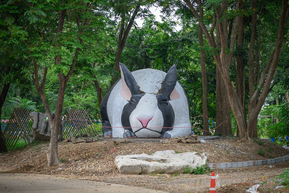 泰國北碧府野生動物園完整攻略|花豹長頸鹿與裝飾景觀玩法解析 10 泰國北碧北碧府野生動物園|綠樹林,大型兔子雕塑,土丘,泥土地面,木棧柵欄,橘白交通錐,戶外園區,閉眼兔頭塑像