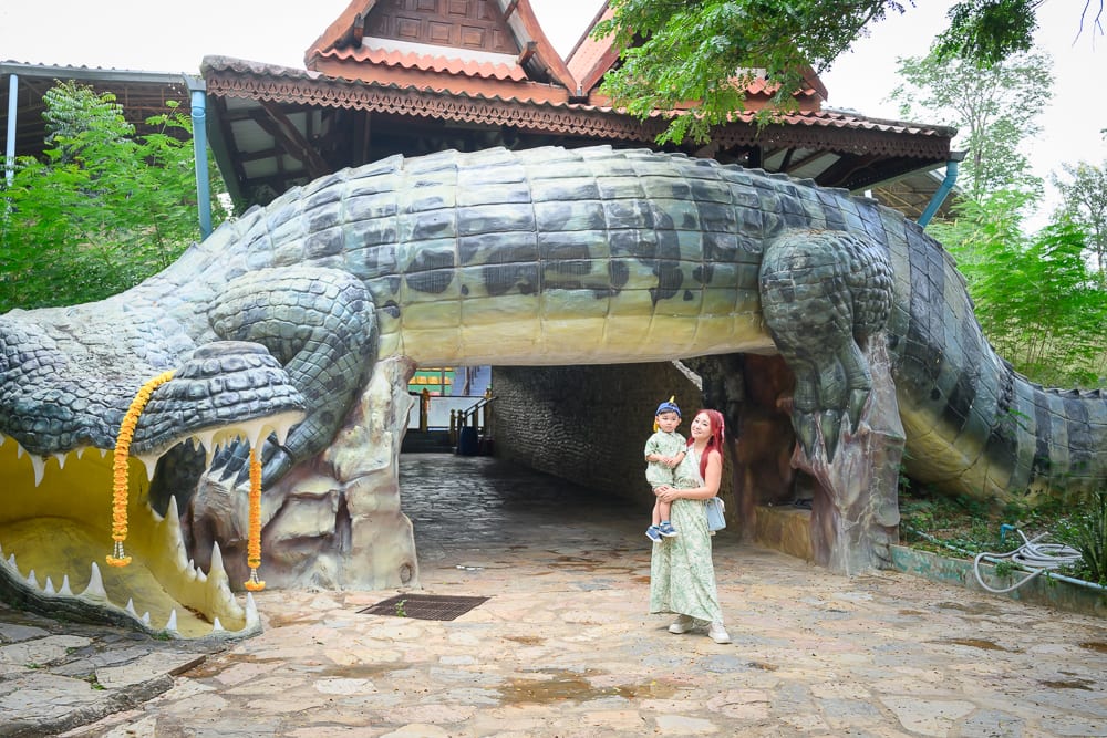 泰國北碧府野生動物園完整攻略|花豹長頸鹿與裝飾景觀玩法解析 8 泰國北碧北碧府野生動物園|女性、小孩、巨型鱷魚造景、寺廟、石板地。紅髮女子站在巨大鱷魚造景的腹部下方,懷中抱著一名戴帽男童,鱷