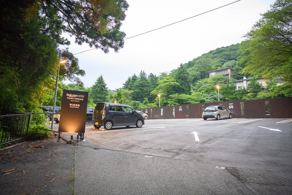 日本箱根-箱根神社｜戶外停車場，黑色休旅車，白色小型車，指引箭頭，立牌，金屬圍籬，綠樹，山坡建築。