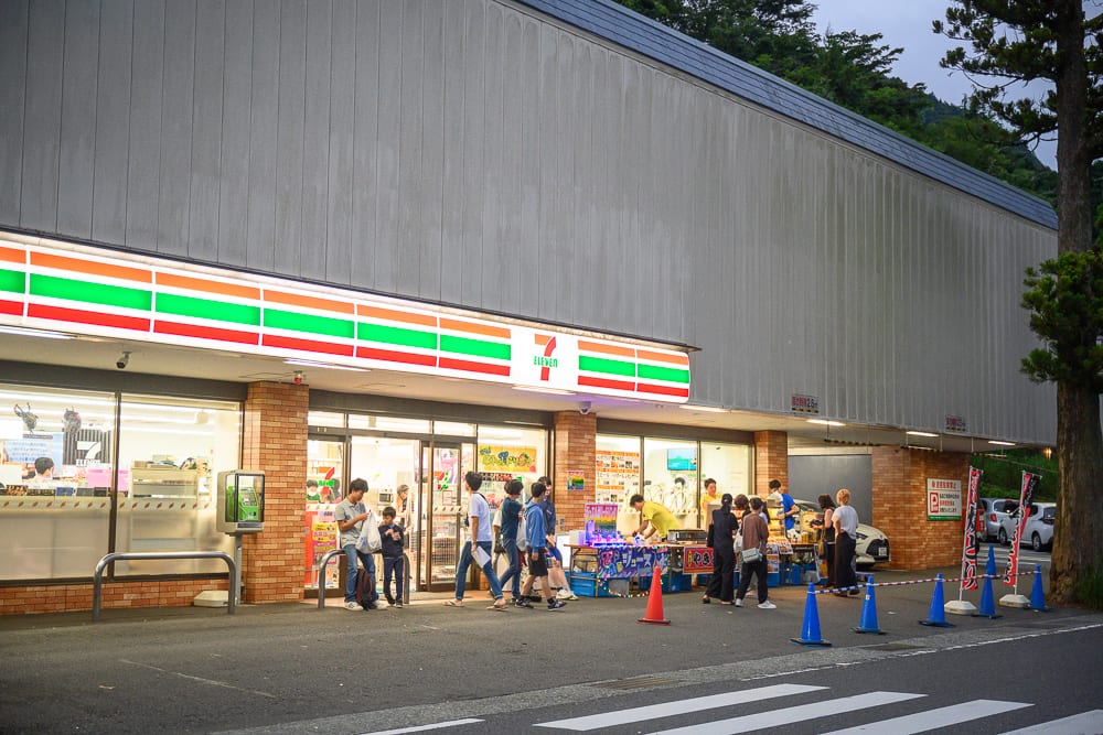 日本箱根-箱根神社｜便利商店，自動門，招牌，人群，街道，三角錐，黃色燈光，灰牆，柏油路，汽車
