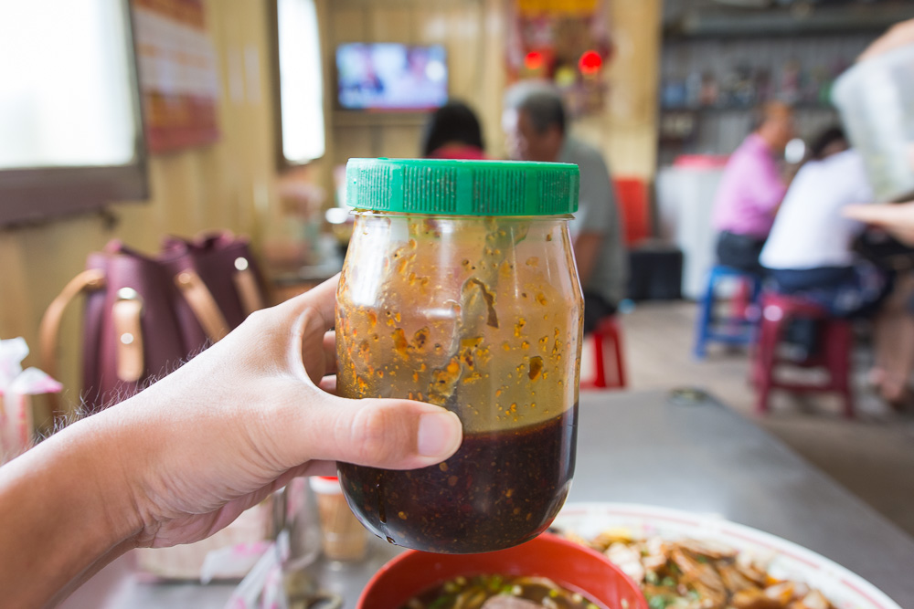 苗栗外省牛肉麵|牛肉麵配酸菜與辣椒油,滷豆干、豬耳朵切盤可當小菜 5 苗栗外省牛肉麵|手持辣椒油罐,背景為室內小吃店,有桌椅與用餐民眾。