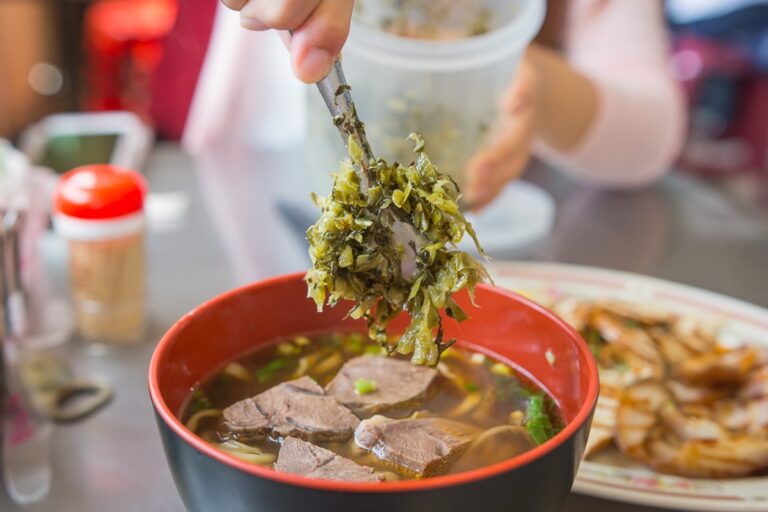 苗栗外省牛肉麵｜餐桌上，手持夾子將酸菜放入牛肉麵碗中，旁有小菜。