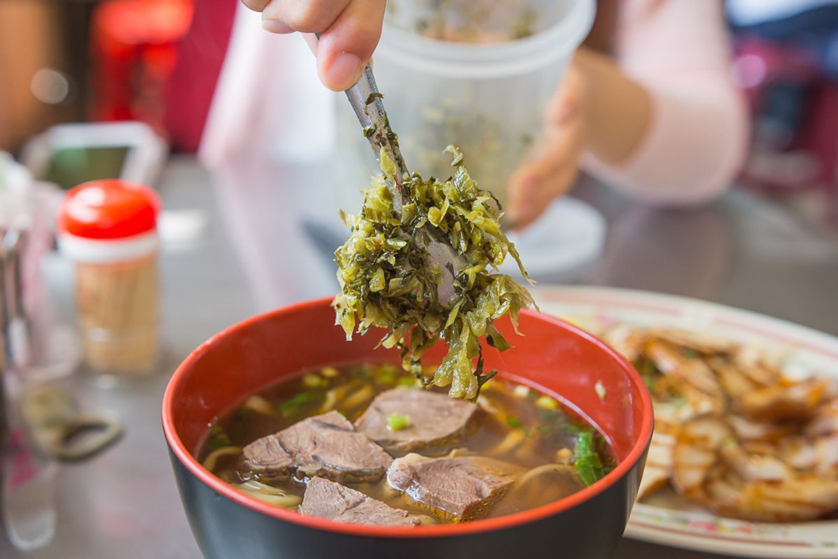 苗栗外省牛肉麵|牛肉麵配酸菜與辣椒油,滷豆干、豬耳朵切盤可當小菜 1 苗栗外省牛肉麵|餐桌上,手持夾子將酸菜放入牛肉麵碗中,旁有小菜。