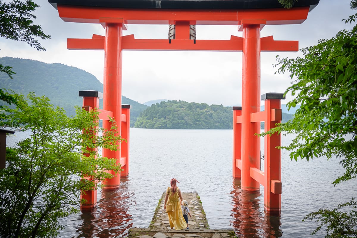 日本箱根-箱根神社｜鳥居、湖、山、石棧道、人物，紅色大型神社鳥居立於湖水中，遠處為群山與水域。石砌棧道延伸至湖面，一名穿黃長裙的成人與一名戴帽小孩背對鏡頭站在棧道上，兩側綠樹枝葉垂掛。