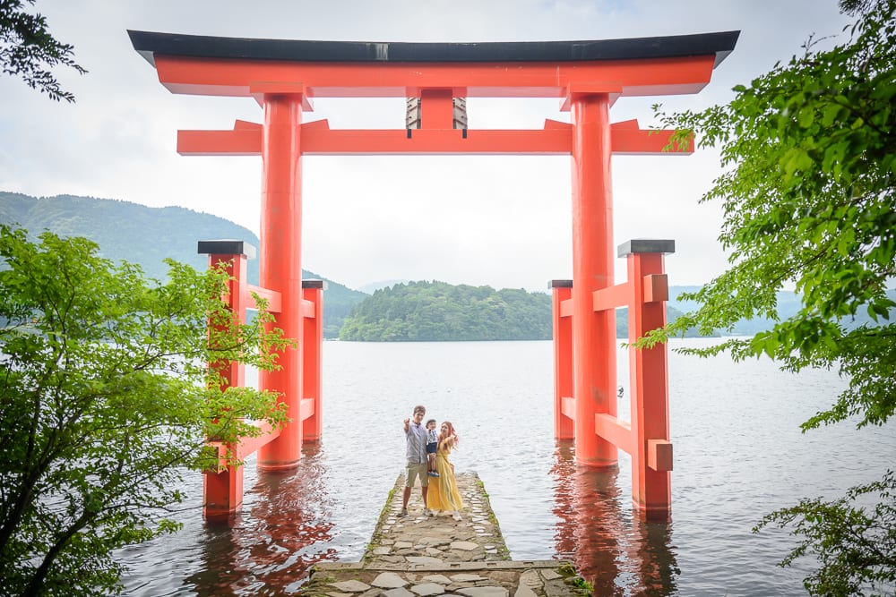 日本箱根-箱根神社｜鳥居、湖、石砌步道、樹木、遠山、三人家庭，灰白雲層下的水面上立著巨大朱紅色鳥居，石板延伸進湖中，兩旁有綠樹枝葉框出畫面。