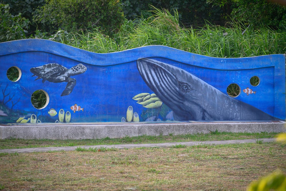 桃園海寧公園兒童遊戲場完整指南|螺旋滑梯與遊具玩法 4 桃園海寧公園|牆壁、藍色、壁畫、海龜、鯨魚、小魚、圓孔、草地。戶外綠地設置波浪造型的藍色水泥牆,牆面彩繪海洋生物,左側有海龜與小丑魚,右側為巨大鯨魚。