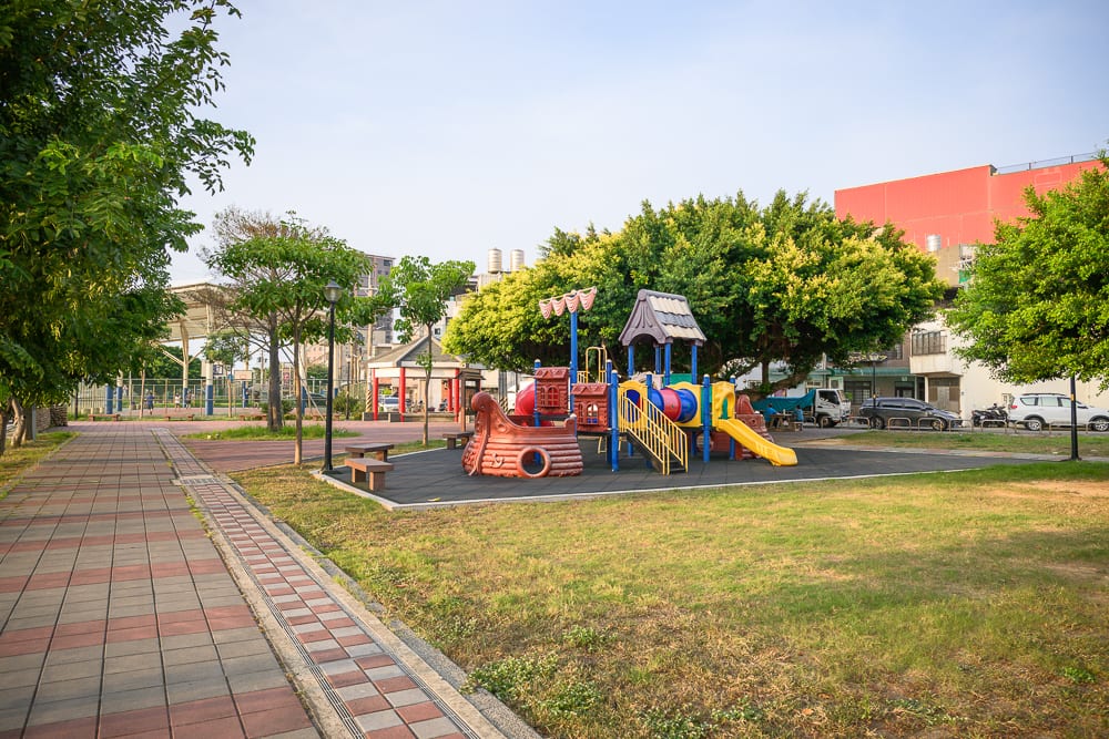 桃園海寧公園兒童遊戲場完整指南|螺旋滑梯與遊具玩法 11 桃園海寧公園|公園、步道、溜滑梯、草皮、大樹,紅磚拼貼地磚一路延伸,彩色塑膠遊具設立於黑色軟墊上,周圍有綠樹環繞與低矮建物。