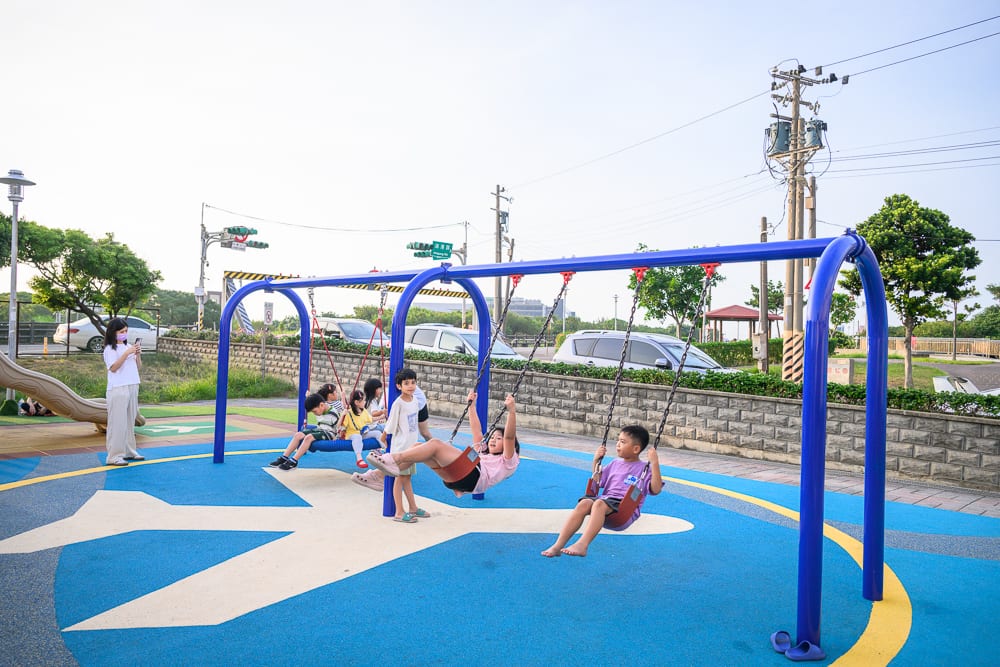 桃園海寧公園兒童遊戲場完整指南|螺旋滑梯與遊具玩法 8 桃園海寧公園|戶外公園,遊樂場,藍色金屬盪鞦韆,多位孩童盪鞦韆,飛機圖案地墊,電線桿,汽車,成人拿手機拍照,綠樹,石牆。