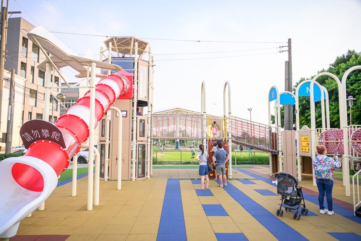桃園海寧公園兒童遊戲場完整指南|螺旋滑梯與遊具玩法 1 桃園海寧公園|遊樂場、溜滑梯、塔樓、繩網、推車、行人,黃藍相間的地墊鋪面,紅色螺旋滑梯延伸至前,兩側有建築物與綠樹。