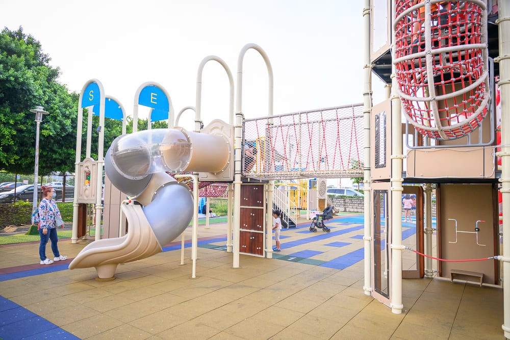 桃園海寧公園兒童遊戲場完整指南|螺旋滑梯與遊具玩法 10 桃園海寧公園|戶外遊樂場,溜滑梯,攀爬網,塑膠管狀滑道,軟墊地面,路燈,綠樹,行人,車輛。