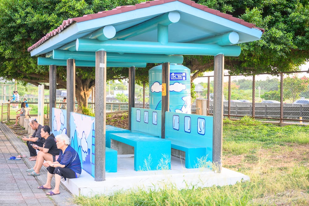 桃園海寧公園兒童遊戲場完整指南|螺旋滑梯與遊具玩法 5 桃園海寧公園|戶外,涼亭,座椅,人群,水塔,樹木,草地,鐵網欄杆。藍色屋頂的涼亭內外設置長椅,數名民眾坐在椅凳上。後方矗立著藍色圓柱水塔與枝葉繁茂的大樹,周圍環繞綠草地與金屬圍欄。