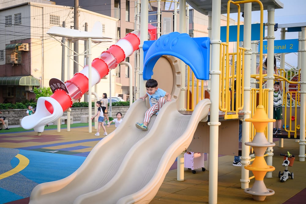 桃園海寧公園兒童遊戲場完整指南|螺旋滑梯與遊具玩法 7 桃園海寧公園|兒童遊戲場,滑梯,兒童,遊樂設施,建築物。小男孩正從雙道滑梯滑下,後方有多段式紅色螺旋滑管,周圍站立著幾位孩童與大人,背景為多層樓住宅。