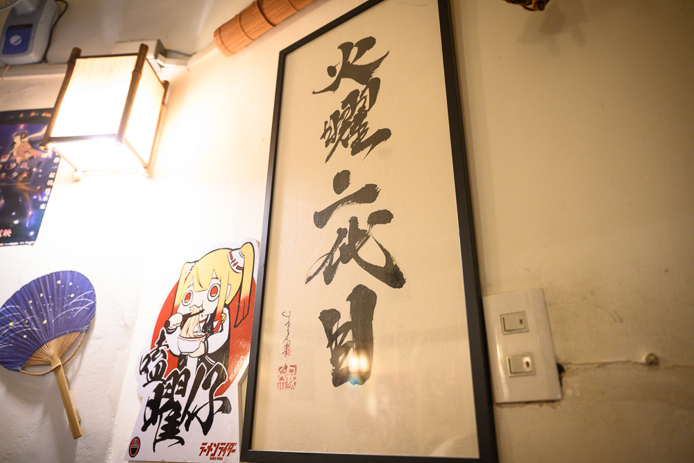 台中火曜二代目拉麵｜牆面掛有書法字畫與動漫貼紙，旁有木框燈具與電燈開關，呈現日式風格室內空間。