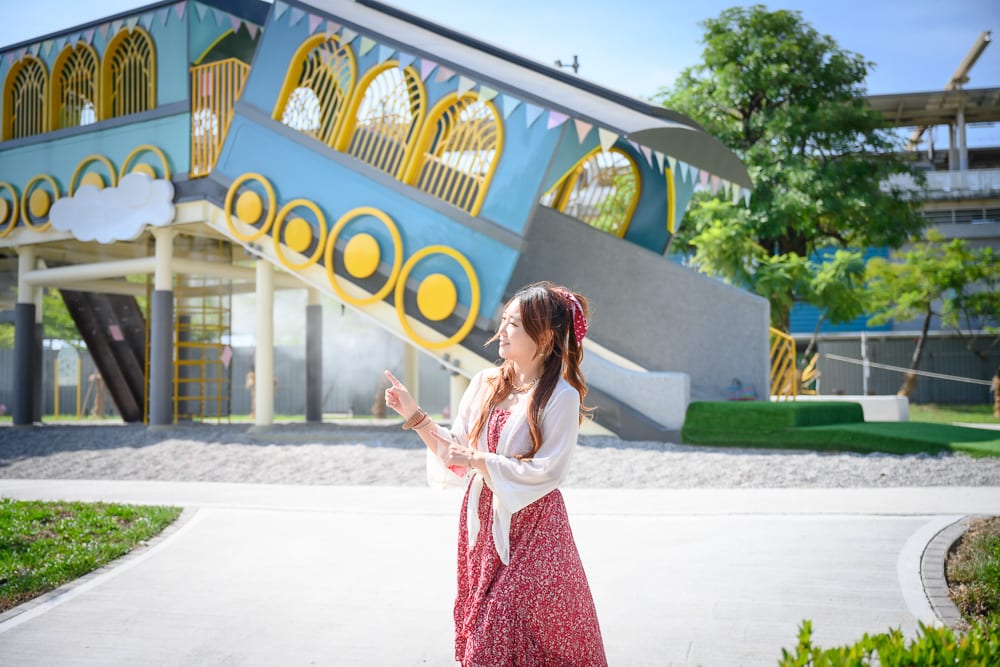 屏東林邊車站公園｜女子、紅洋裝、戶外公園、藍色火車造型遊樂設施、溜滑梯、水泥地、綠樹、藍天白雲。女子站立於白色步道上，側身抬手指向後方建物。