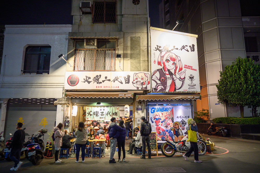 台中火曜二代目拉麵｜夜晚的拉麵店，招牌繪有動漫角色，店外停放機車，顧客在騎樓用餐。