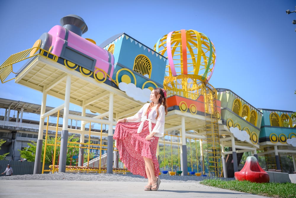 屏東林邊車站公園｜女子、火車裝置藝術、熱氣球鋼架、戶外遊樂場、藍天，粉藍相間的巨大列車模型架高於水泥地，女子身穿紅底碎花長裙與白色罩衫，側身站立且手提裙擺。