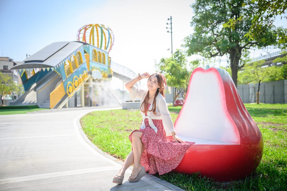 屏東林邊車站公園｜女子，紅裙，涼鞋，蓮霧椅，草地，步道，遊樂場，溜滑梯。女子側身坐在紅色的蓮霧造型長椅上，身穿碎花洋裝與白色罩衫，左手撐在椅背，雙腿交疊看向鏡頭。背景為戶外公園，左側有大型彩色遊樂設施與階梯，右側有綠樹與草皮，水泥步道貫穿畫面。