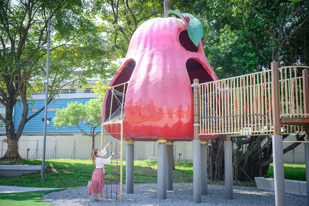 屏東林邊車站公園｜戶外公園、巨型紅色水果造型溜滑梯、攀爬網、木棧道、女性、綠樹、草地，穿著粉色長裙的女子正拉著攀爬網繩索，周圍有金屬欄杆與路燈。