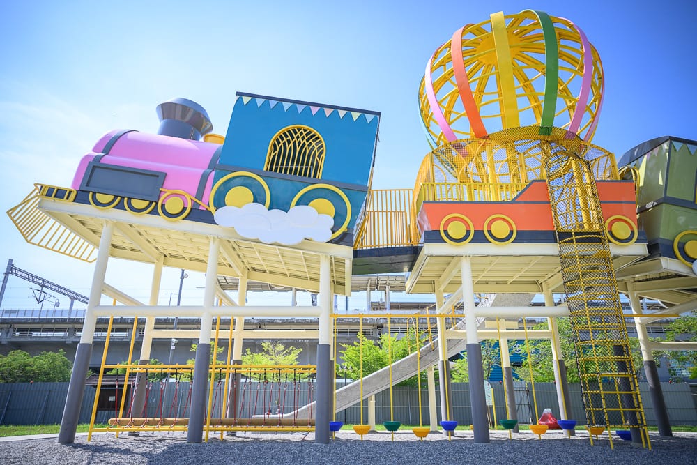 屏東林邊車站公園｜高架平台、火車造型遊具、熱氣球骨架、攀爬網、滑梯、支柱、藍天。色彩繽紛的火車與熱氣球大型遊具架高設置於金屬支柱上，下方鋪面碎石，背景為藍天與灰色結構。