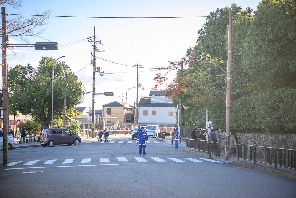 日本京都金閣寺完整指南|池畔倒影與庭園步道 7 日本京都金閣寺|街道,路口,斑馬線,交管人員,車輛,電線桿,房屋,綠樹。交通路口處,穿著藍色制服的交管人員站在斑馬線中央,四周有停放的汽車與行走民眾。